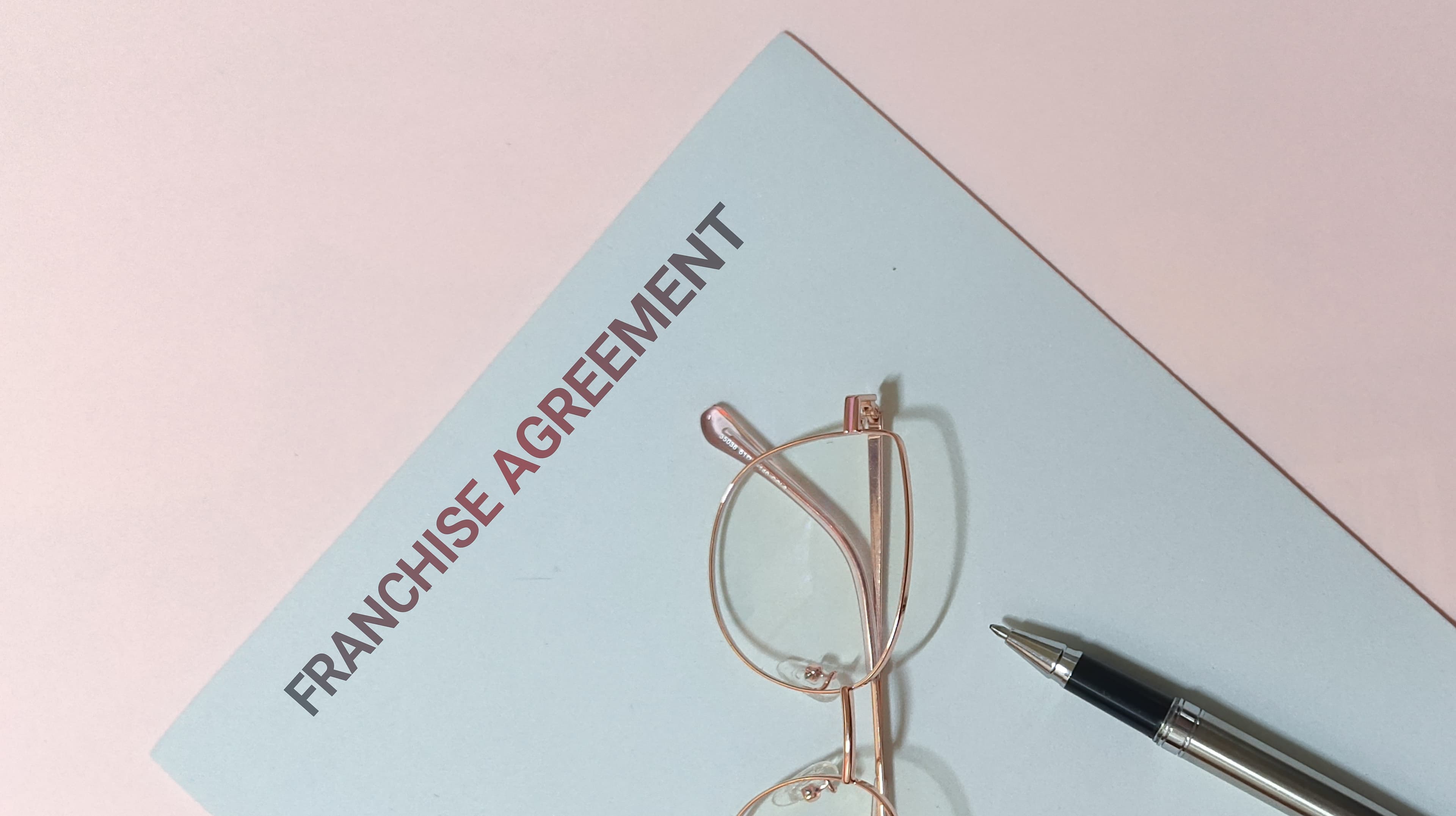 Franchise agreement document, rose gold glasses, and a pen on a light pink background.