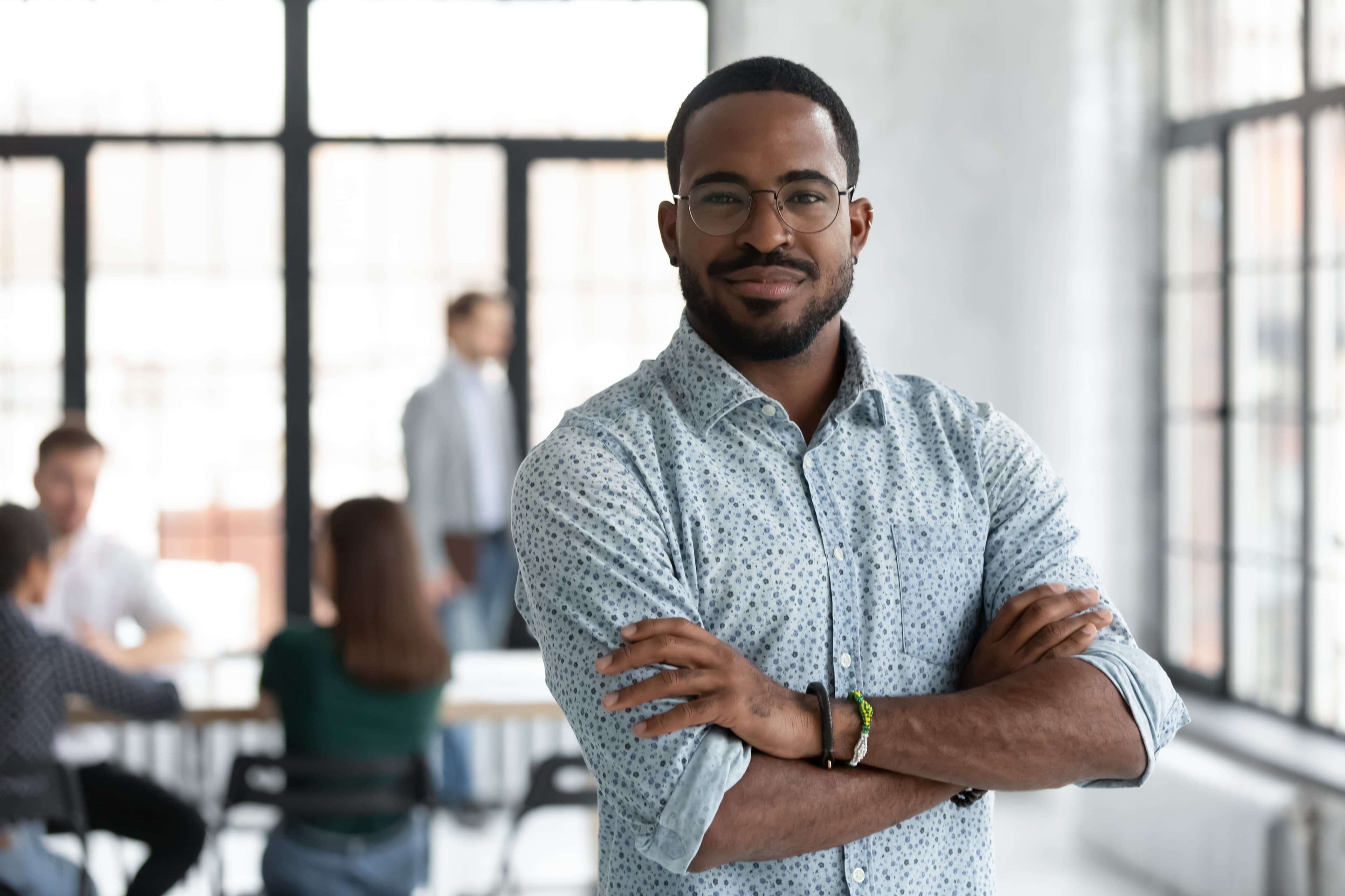 Confident Black professional with glasses and crossed arms in a bright, modern office environment.
